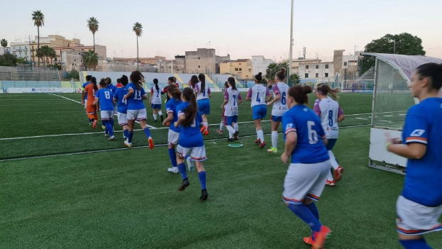 Eine Gruppe von Frauen, die auf einem Feld mit Gebäuden, Bäumen und Pfählen umgeben Fußball spielen, unter einem klaren blauen Himmel, mit einem Torpfosten und Netz auf der rechten Seite.