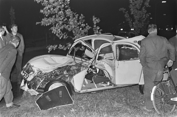 Schwarzes und weißes Bild eines verunglückten Autos am Straßenrand mit Menschen und einem Fahrrad in der Nähe, Bäume und beleuchtete Lichter im Hintergrund.