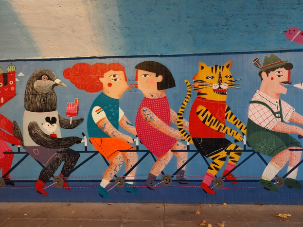 Mural an einer Gebäudeseite, das Menschen auf Fahrrädern auf einer Straße mit einem Himmelhintergrund zeigt, Teil einer Londoner Street-Art-Serie.