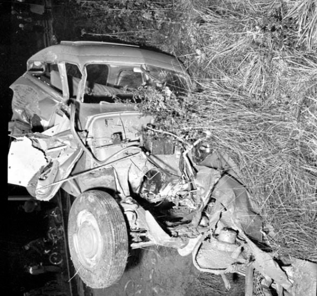 Ein Schwarz-Wei-Foto eines schwer beschädigten Autos am Straßenrand, mit einer zerknärzten Front, zerbrochenen Fenstern, einem Baum im Hintergrund und verstreuten Gegenständen um die Unfallstelle.