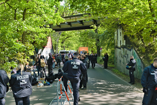 Polizeibeamte hinter einer Barriere auf einer Straße stehend mit Menschen in der Nähe, Bäume und eine Brücke im Hintergrund und eine Wand mit Schrift auf der rechten Seite.