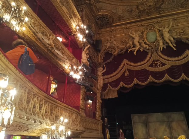 Gemälde namens "Tanz am Moulin Rouge" von Edgar Degas zeigt tanzende Frauen in weißen Kleidern und bunten Kostümen auf einer Bühne mit einem lebhaften blauen Hintergrund und warmem gelbem Licht.