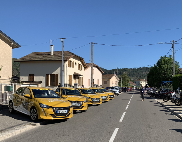 Eine Reihe gelber Taxis, die entlang einer Straße geparkt sind, mit Fußgängern auf dem benachbarten Gehweg, vor einem Hintergrund aus Gebäuden, Bäumen, Strommasten und einem klaren blauen Himmel.