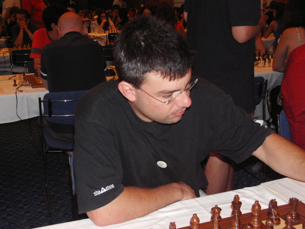 Ein Mann in einem schwarzen T-Shirt spielt Schach an einem Tisch, im Hintergrund spielen Gruppen von Menschen Schach und es gibt eine blaue Matte auf dem Boden.