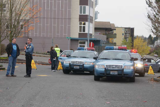 Autos auf einer Straße mit vier Menschen in der Nähe, Gebäuden mit Fenstern im Hintergrund, Bäumen und Warn Dreiecken.