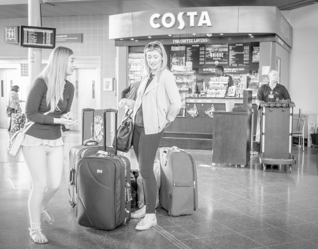 Zwei Frauen vor mehreren Gepäckstücken mit einem Kaffeeschop im Hintergrund, wahrscheinlich an einem Flughafen.
