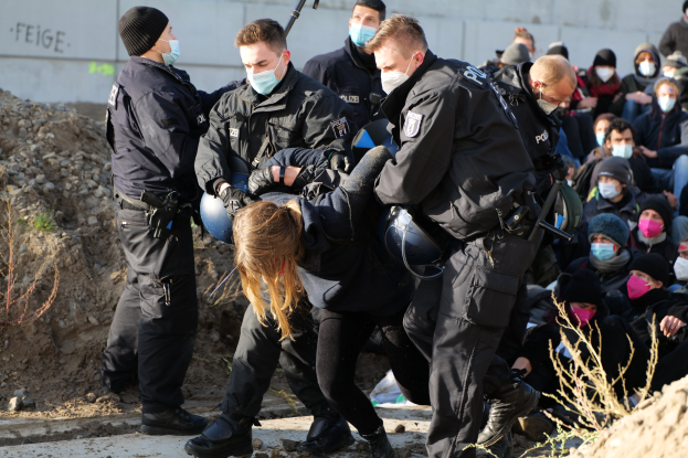 Gruppe maskierter Personen in Mützen, einige bewaffnet, umringt eine Frau, die von der Polizei eskortiert wird, in einer städtischen Umgebung mit Pflanzen, Steinen und protestbezogenen Wandtexten im Hintergrund.