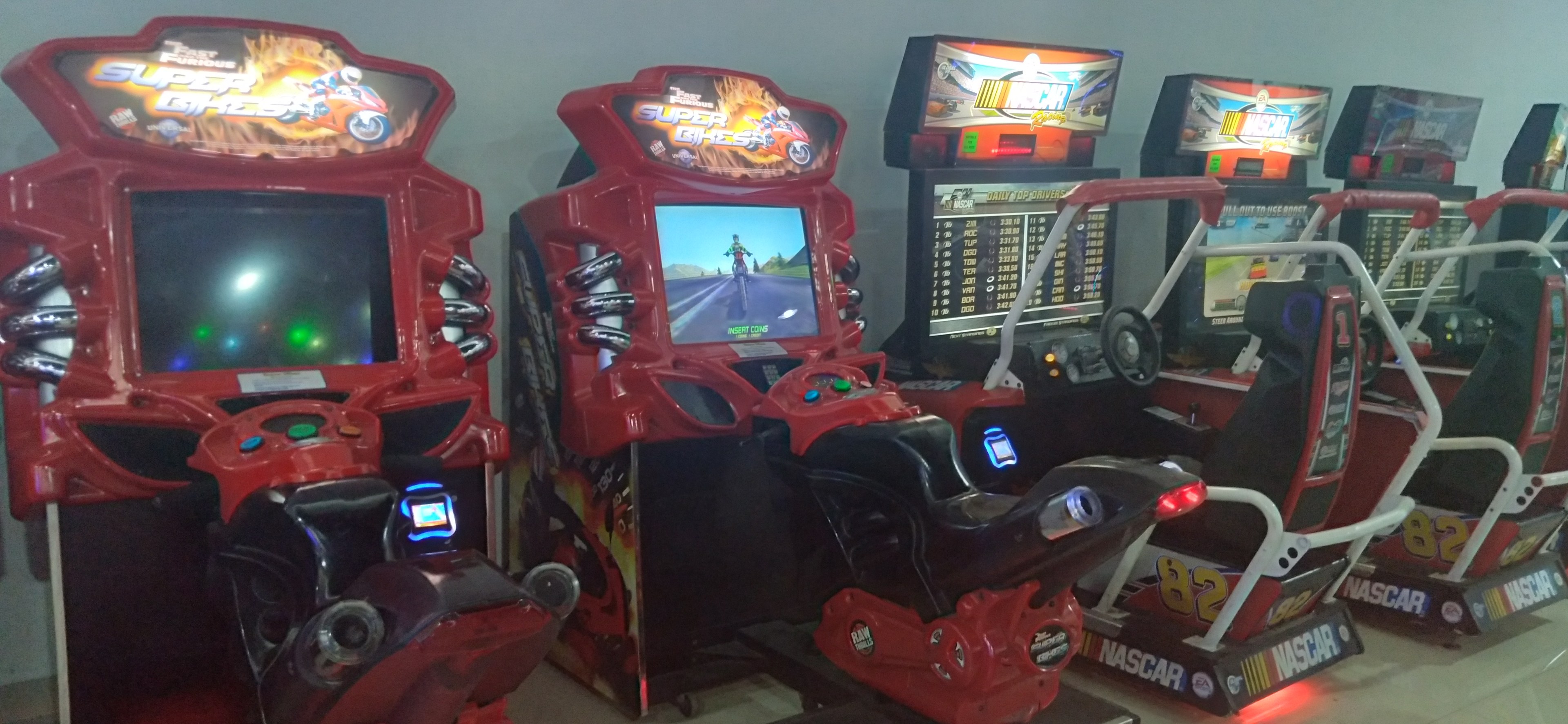 Eine Reihe buntfarbiger Arcade-Rennwagenautomaten auf einem gefliesten Boden mit einer Wand im Hintergrund.