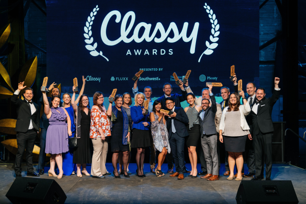 Eine Gruppe von Menschen auf einer Bühne, die lächeln und Auszeichnungen halten, mit Lautsprechern unten und einem Hintergrundbild mit der Aufschrift 'Classy Awards 2019'.
