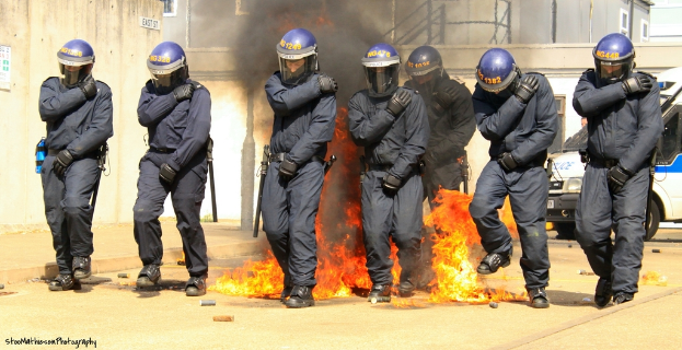 Menschen in Helmen stehen vor einem Feuer mit Gegenständen auf dem Boden, Gebäuden im Hintergrund, einem Fahrzeug, einem Plakat und einer Tafel an der linken Wand und Text unten.