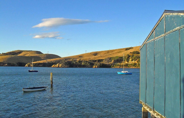 Boote und Pfähle im Wasser, ein hölzernes Haus rechts, mit Bäumen und Bergen mit Windmühlen im Hintergrund, unter einem bewölkten Himmel.