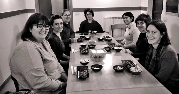 Eine Gruppe von sieben Frauen sitzt an einem Tisch mit Essen, Tassen, Tellern und einem Glas tragbaren Gerät in der linken Ecke.