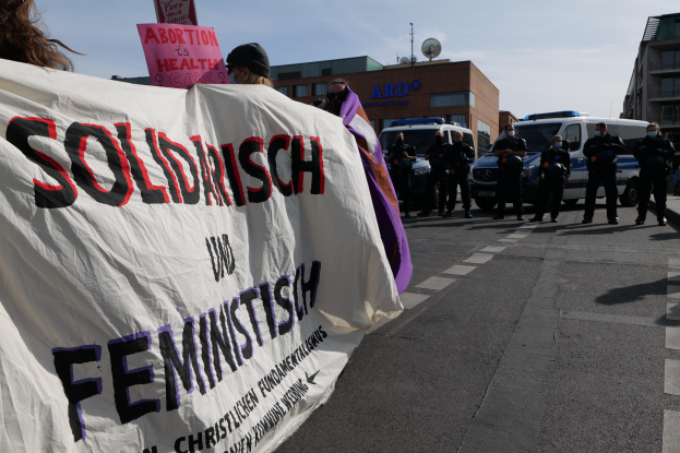 Eine Gruppe von Menschen marschiert mit einem Banner, auf dem 'Solidarität und Feminismus' steht, eine Straße entlang, mit parkenden Fahrzeugen und Gebäuden im Hintergrund bei klarem blauem Himmel.