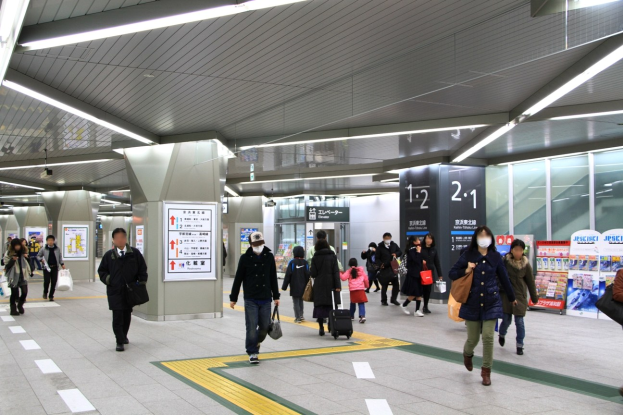 Menschen mit Gepäck durch einen Bahnhof gehen und übergeordnete Schilder mit Text und Zahlen anzeigen.