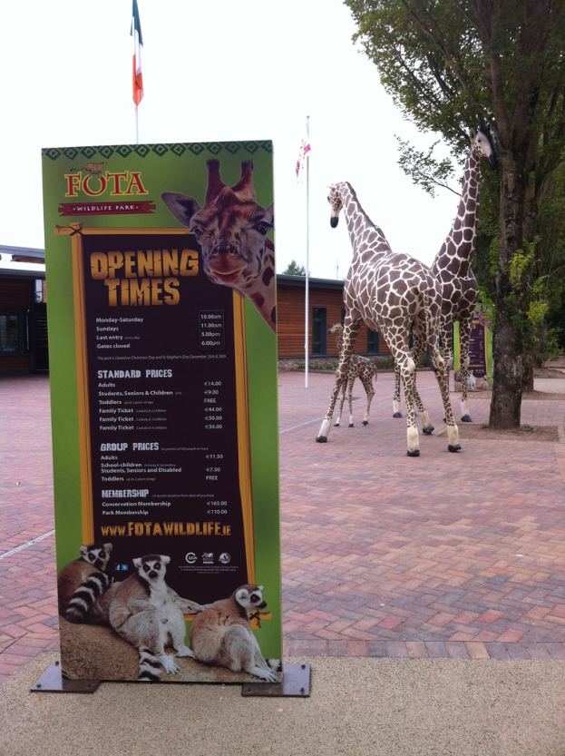 Giraffen stehen neben Bäumen, Schuppen und Flaggen, mit einer Tafel, die Text, Logos und Tierbilder anzeigt.