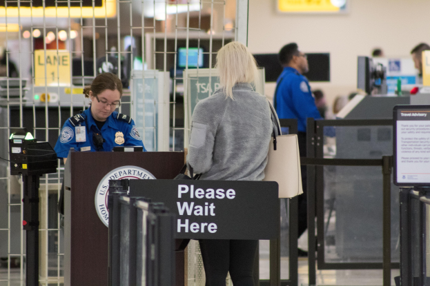 Eine Frau in einer grauen Jacke und schwarzen Hosen steht an einem Flughafen-Metallsensor und hält eine Handtasche, mit einem Polizeibeamten in einer blauen Uniform zu ihrer Linken und einer Tafel dahinter.