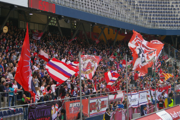 Eine große Menschenmenge in einem Stadion schwenkt Fahnen und Banner mit Text, umgeben von Geländern, mit einer digitalen Anzeigetafel oben sichtbar.