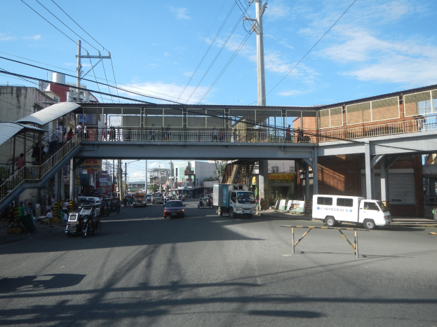 Stadtstraße mit fahrenden Fahrzeugen, eine Fußgängerbrücke mit Geländern und Stufen, Menschen auf der Brücke, Strommasten mit Drähten, Gebäude mit Fenstern und ein bewölkter Himmel im Hintergrund.