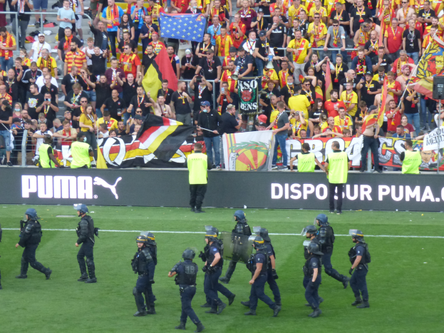 Eine Gruppe von Polizisten in Helmen und mit Waffen in der Hand geht über ein Fußballfeld, im Hintergrund sind Menschen mit Fahnen und Schildern sowie Absperrungen zu sehen.