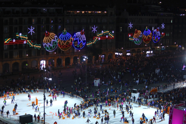 Eine große Menschenmenge, die auf einer Eisbahn vor einem Gebäude Schlittschuh läuft, beleuchtet von Straßenlaternen und geschmückt mit festlichen Weihnachtsdekorationen, umgeben von Geländern und Pfosten und einem dunklen Hintergrund.