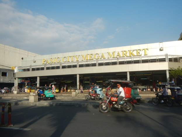 Eine belebte Straßenszene auf dem Pasig City Mega Market mit Fahrzeugen und Fußgängern, ein Gebäude mit Text im Hintergrund und ein bewölkter Himmel.
