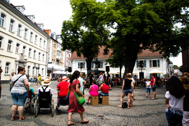 Eine Gruppe von Menschen, einige im Rollstuhl, geht und sitzt auf einem Kopfsteinpflasterweg in der Altstadt von Heidelberg, mit Bäumen, Gebäuden, Laternen und einer Statue im Hintergrund unter einem bewölkten Himmel.