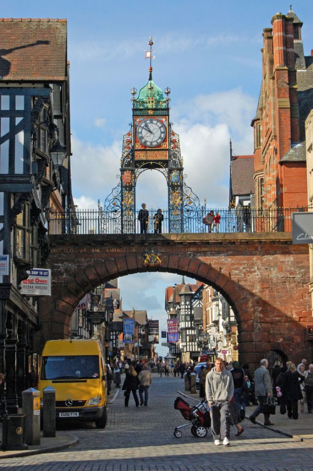Eine belebte Straße mit Fußgängern, ein gelber Lieferwagen auf dem linken Gehweg, eine Brücke mit zwei Personen darüber, eine Uhr über der Brücke, Gebäude auf beiden Seiten und der Himmel darüber.