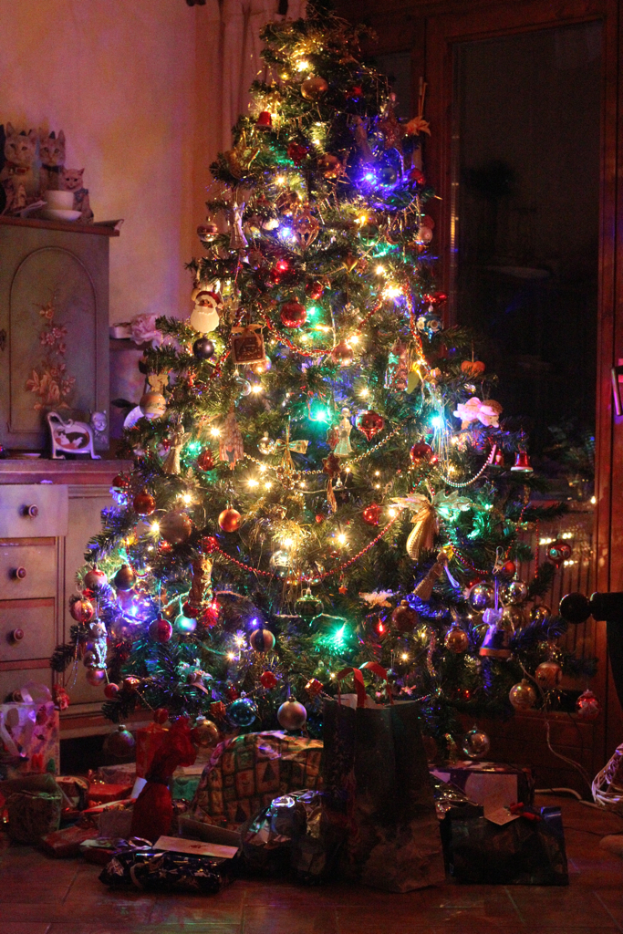 Ein Weihnachtsbaum mit Lichtern und Dekorationen, Geschenke auf dem Boden, ein Schrank mit Gegenständen auf der linken Seite und ein Fenster an der rechten Wand.