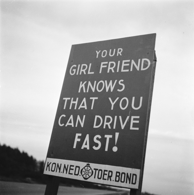 Ein schwarzes und weißes Schild mit der Aufschrift "Deine Freundin weißt, dass du schnell fahren kannst" umgeben von Bäumen und einem klaren Himmel.