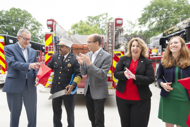 Gruppe von Menschen, die bei einer Eröffnungszeremonie der Alexandria Fire Department klatschen und lächeln, mit zwei Personen, die eine Schere und ein rotes Band halten, vor einem Feuerwehrauto, Bäumen, Drähten und einem klaren blauen Himmel im Hintergrund.