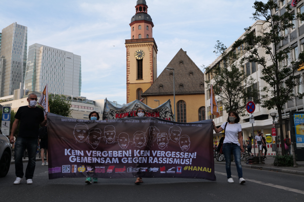 Eine Gruppe von Menschen in Masken, die eine Plakattafel tragen, geht eine Straße entlang, mit einem geparkten Auto auf der linken Seite, Gebäuden und Bäumen im Hintergrund und einem klaren blauen Himmel darüber.