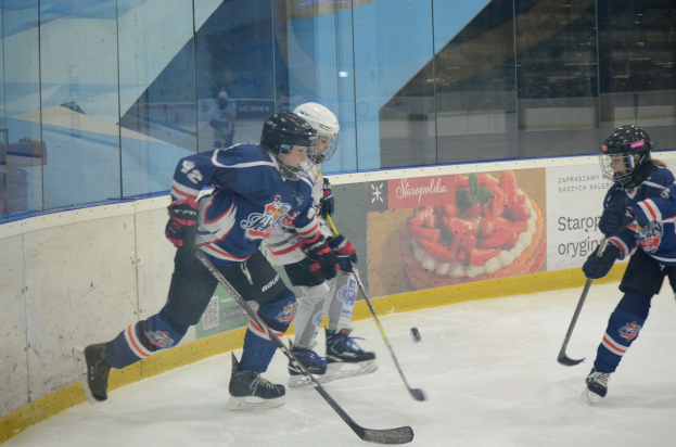 Gruppe junger Menschen, die Eis-Hockey auf einer Indoor-Eisfläche spielen, mit Helmen, Sportuniformen und Schlittschuhen, während sie Hockey-Schläger halten, mit einer Glaswand und einem Plakat im Hintergrund.