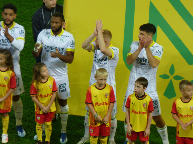 Eine Gruppe junger Menschen in Fußballtrikots steht auf einem Fußballfeld und klatscht feierlich mit einer 'Ligue 1'-Platte im Hintergrund.