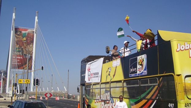 Ein Bus mit Menschen, die Fahnen schwenken, ein Auto links daneben, eine Werbetafel im Hintergrund und Masten mit Verkehrszeichen unter dem Himmel.