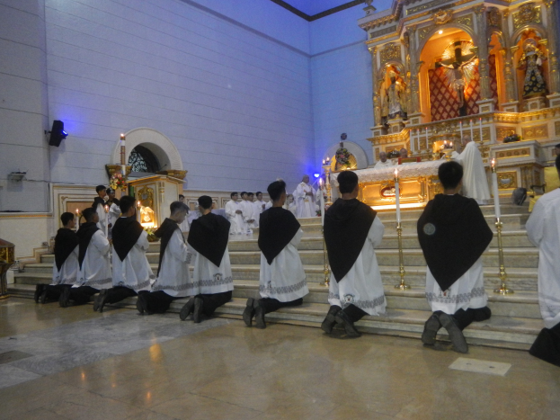 Eine Gruppe von Menschen sitzt auf den Stufen der Kirche mit angezündeten Kerzen vor sich, ein Altar mit Statuen und Gegenständen im Hintergrund und Lautsprecher an der linken Wand angebracht.