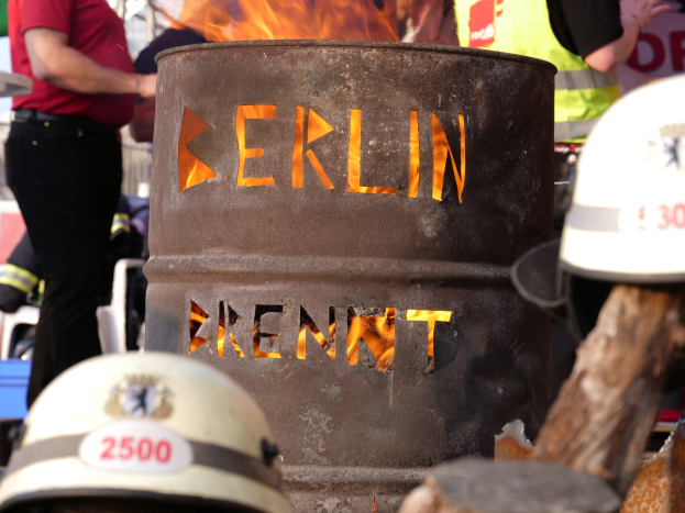 Eine Gruppe von Menschen steht in der Nähe eines Feuerhydranten mit der Aufschrift "Berlin", im Vordergrund ein brennender Eimer, Helme unten und ein hölzernes Objekt rechts.