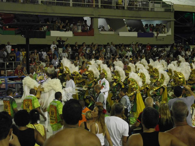 Eine große Menschenmenge, einige in Kostümen, steht vor einem Gebäude bei einem Karneval in Rio de Janeiro, mit Geländern, Pfählen und Lichtern im Hintergrund und einigen Individuen mit Kameras.