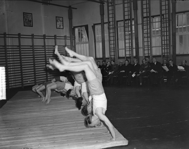 Eine Gruppe von Menschen führt Akrobatik auf einer Bühne vor einem sitzenden Publikum auf, mit Foto-Rahmen, Fenstern und Leitern im Hintergrund, dargestellt in Schwarz-Weiß.