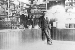 Drei Männer stehen an einem Tresen in einem Laden, umgeben von verschiedenen Gegenständen, mit einem Regal voller Bücher und Gegenständen im Hintergrund, scheinbar an einem Raub beteiligt.