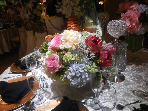 Formaler Esstisch mit Tellern, Besteck, Gläsern, Servietten und einem Blumenstrauß als Tischschmuck, umgeben von Stühlen und im Hintergrund besetzte Tische.