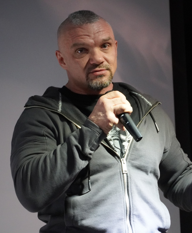 Ein Mann in einer schwarzen Kapuzenjacke und einem schwarzen Anzug mit einem wei├čen Hemd, der ein Mikrofon h├Ąlt und selbstbewusst vor einer Wand steht.