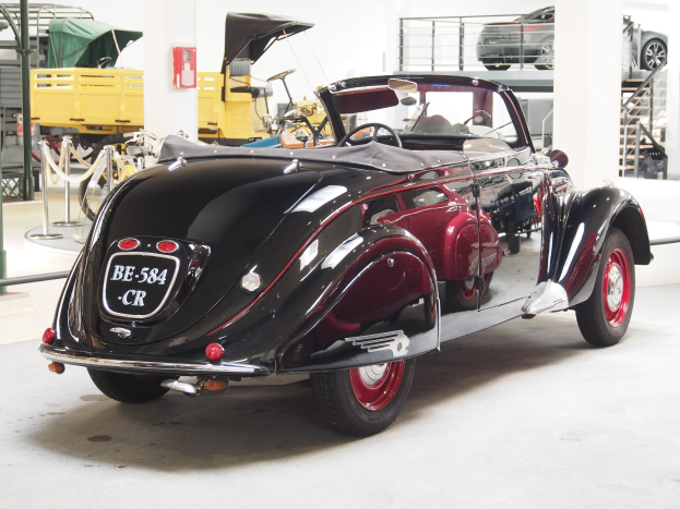 Ein schwarzer und roter Wagen in einer Garage mit anderen Fahrzeugen, Pfählen, Ketten und Gegenständen geparkt, mit einem Feuerlöscher an der Wand im Hintergrund.