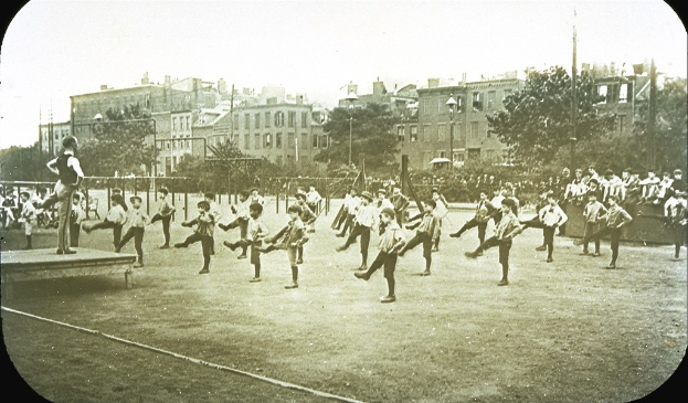 Schwarzes Bild von Kindern, die auf einem Feld mit Baumen, Pfosten und Gebauden im Hintergrund Fuball spielen.