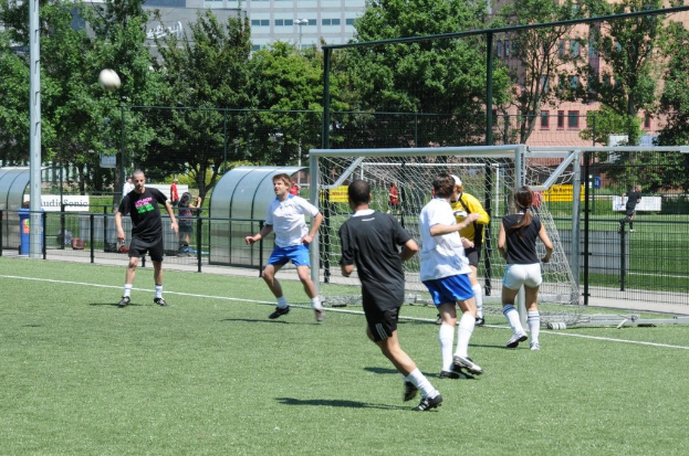 Menschen in Kleidung beim Füßballspielen auf einem Feld mit einem Zaun und einem Tor in der Mitte, einem Pfahl und einem Füßball oben links und Bäumen und Gebäuden im Hintergrund.