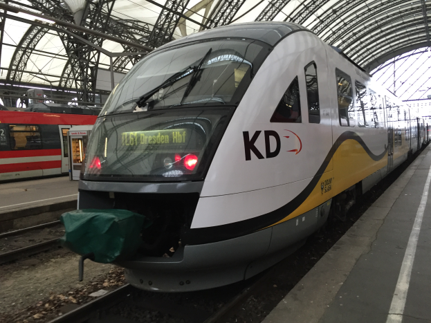 Ein weißer und gelber Zug hält an einem Bahnhof mit einer Plattform auf der rechten Seite, ein weiterer Zug im Hintergrund und Metallstangen und ein Schuppen oben.