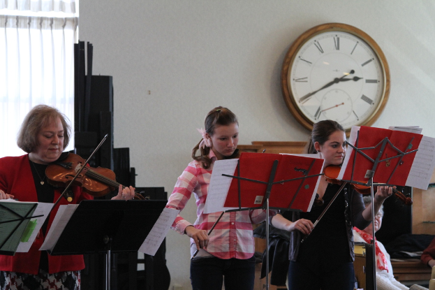 Drei Frauen, die Geigen auf der Bühne spielen, mit Notenpulten und Noten davor, während ein Zuschauer zuhört; eine Uhr ist an der weißen Wand im Hintergrund angebracht, und ein weißer Vorhang ist links sichtbar.