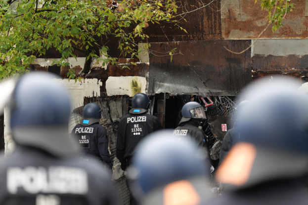 Gruppe von Polizisten in schwarzen Uniformen und Helmen, die vor einem mit Maschendrahtzaun umgebenen Gebäude stehen, mit Bäumen im Hintergrund.
