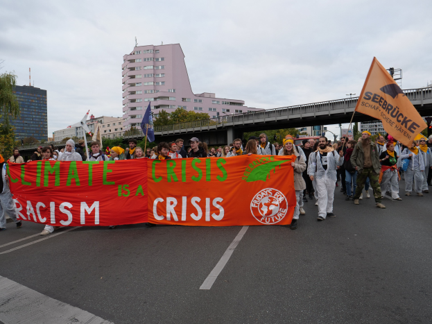 Eine Gruppe von Menschen marschiert eine von Bäumen gesäumte Straße entlang und hält ein Banner hoch, auf dem "Klima-Krise ist eine Krise" steht, mit Gebäuden und einer Brücke im Hintergrund unter einem bewölkten Himmel.