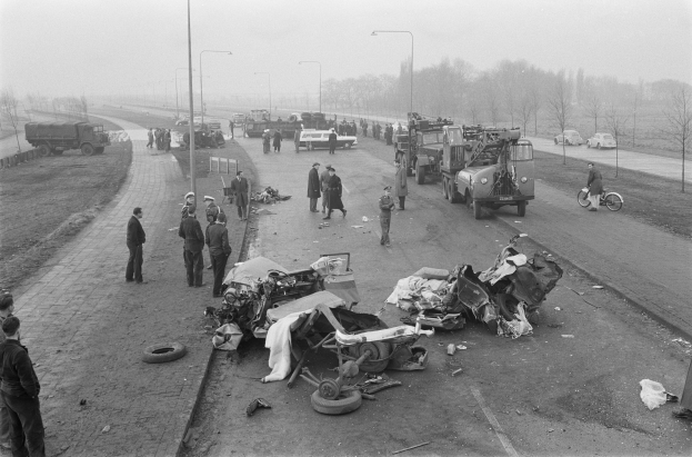 Schwarzes und weißes Bild eines Autounfalls am Straßenrand mit mehreren Fahrzeugen und einer Gruppe von Menschen in der Nähe, Laternenpfähle, Bäume und Himmel im Hintergrund.