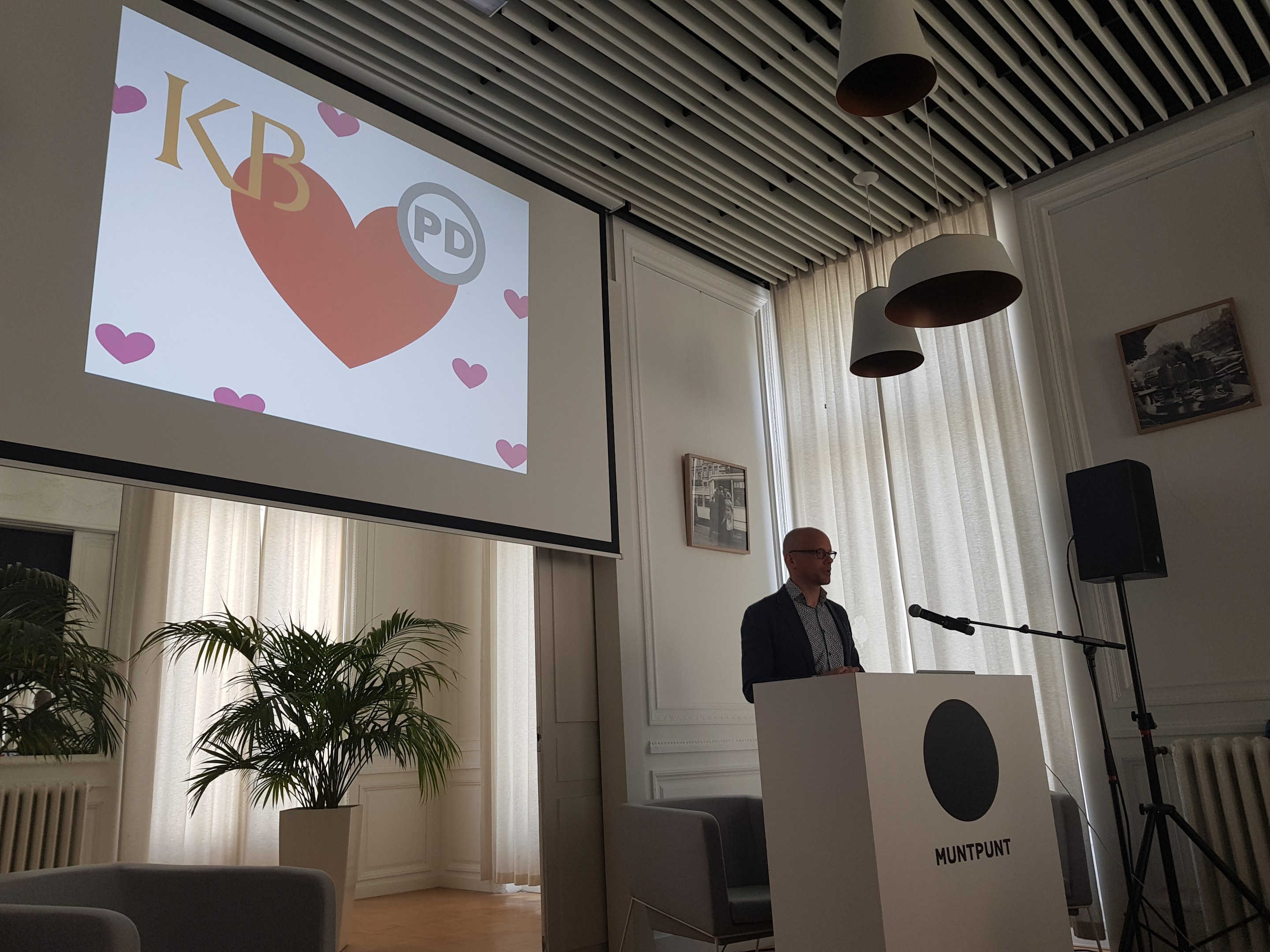 Ein Mann steht an einem Podium mit einem Mikrofon und hält eine Präsentation zum Thema Geschlechtergerechtigkeit, mit einem Projektionsschirm, einem Lautsprecher, Stühlen, einer Topfpflanze, gerahmten Fotos an der Wand, Vorhängen und Deckenleuchten im Raum.